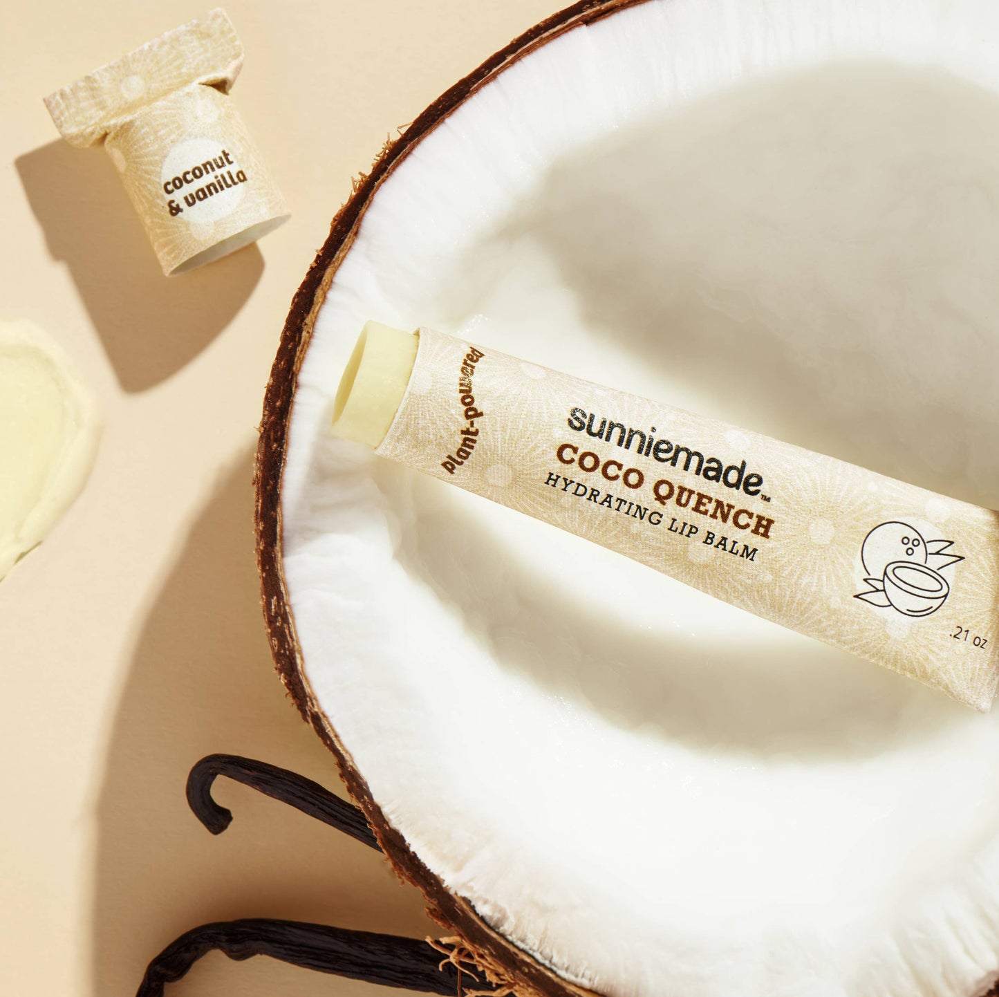 A renewable paper lip balm tube, labeled “Coco Quench Hydrating Lip Balm”, resting inside of a coconut half. The lid is removed, exposing the lip balm itself.