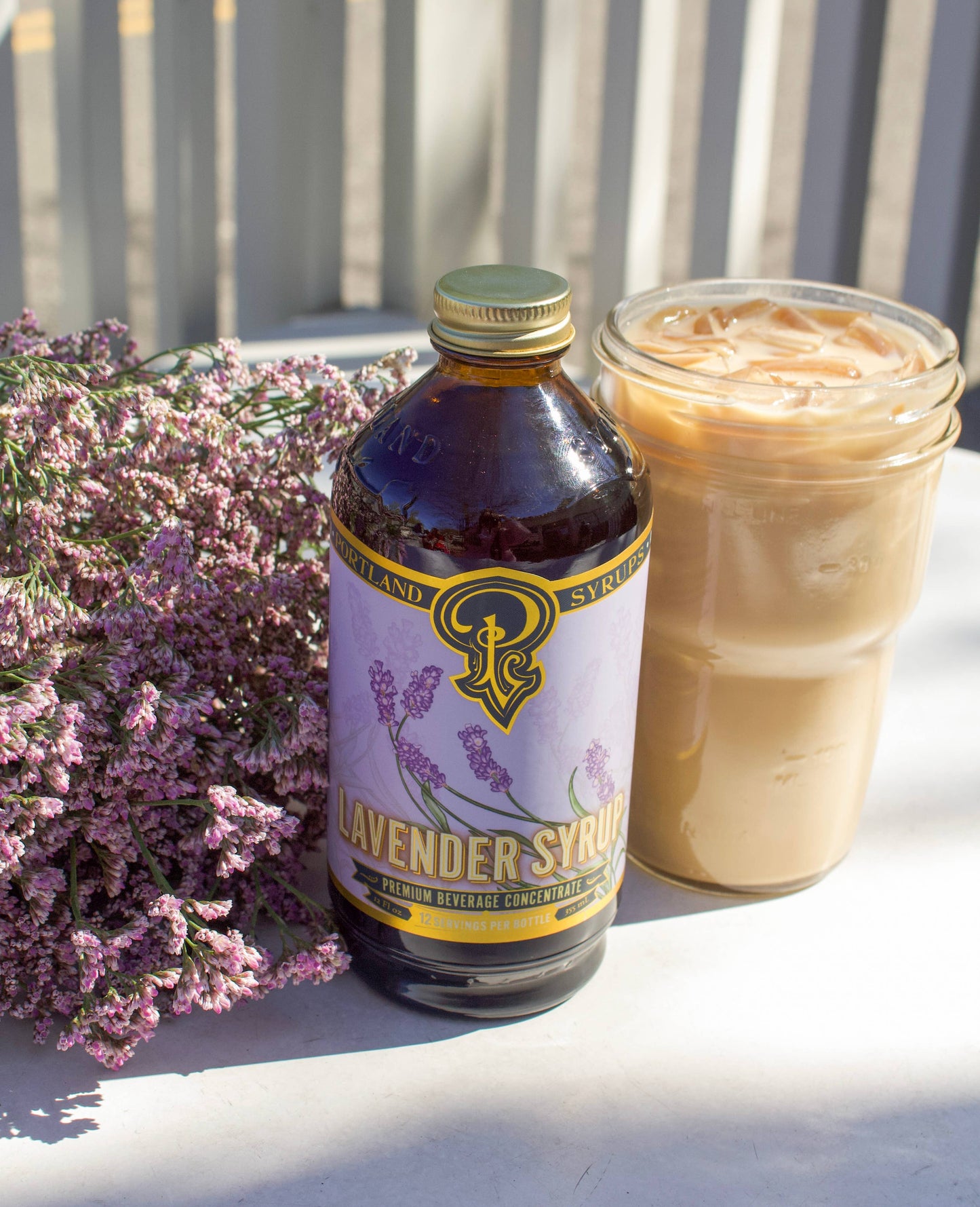 A dark glass bottle labeled “Portland Syrups - Lavender Syrup - Premium Beverage Concentrate”. It sits out in the sunlight next to a glass of iced coffee and a bundle of lavender flowers.