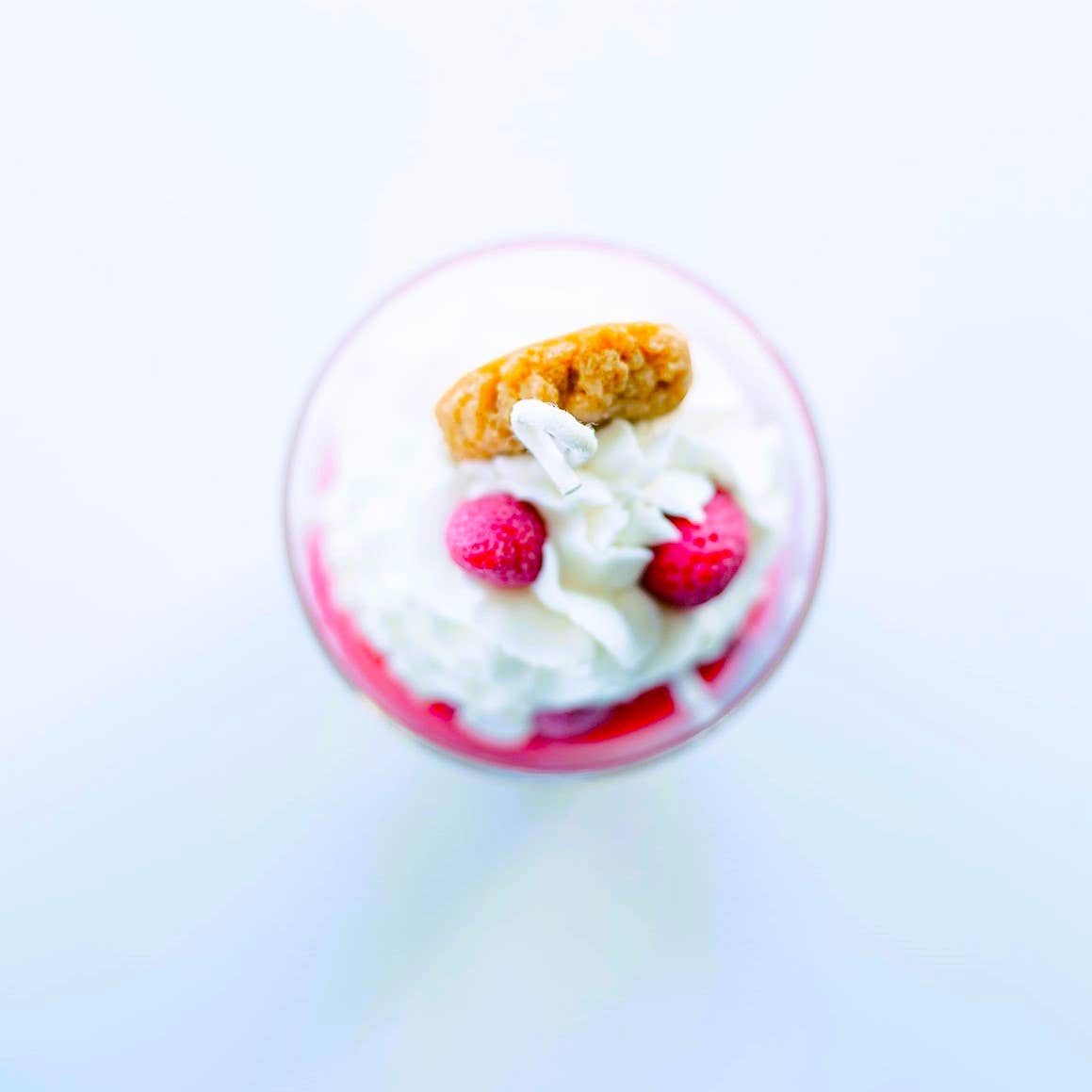 Top-down image of a candle in a clear glass jar, clearly showing that it is topped with decorations of whipped cream, strawberries, and a wafer cookie, all sculpted from wax.
