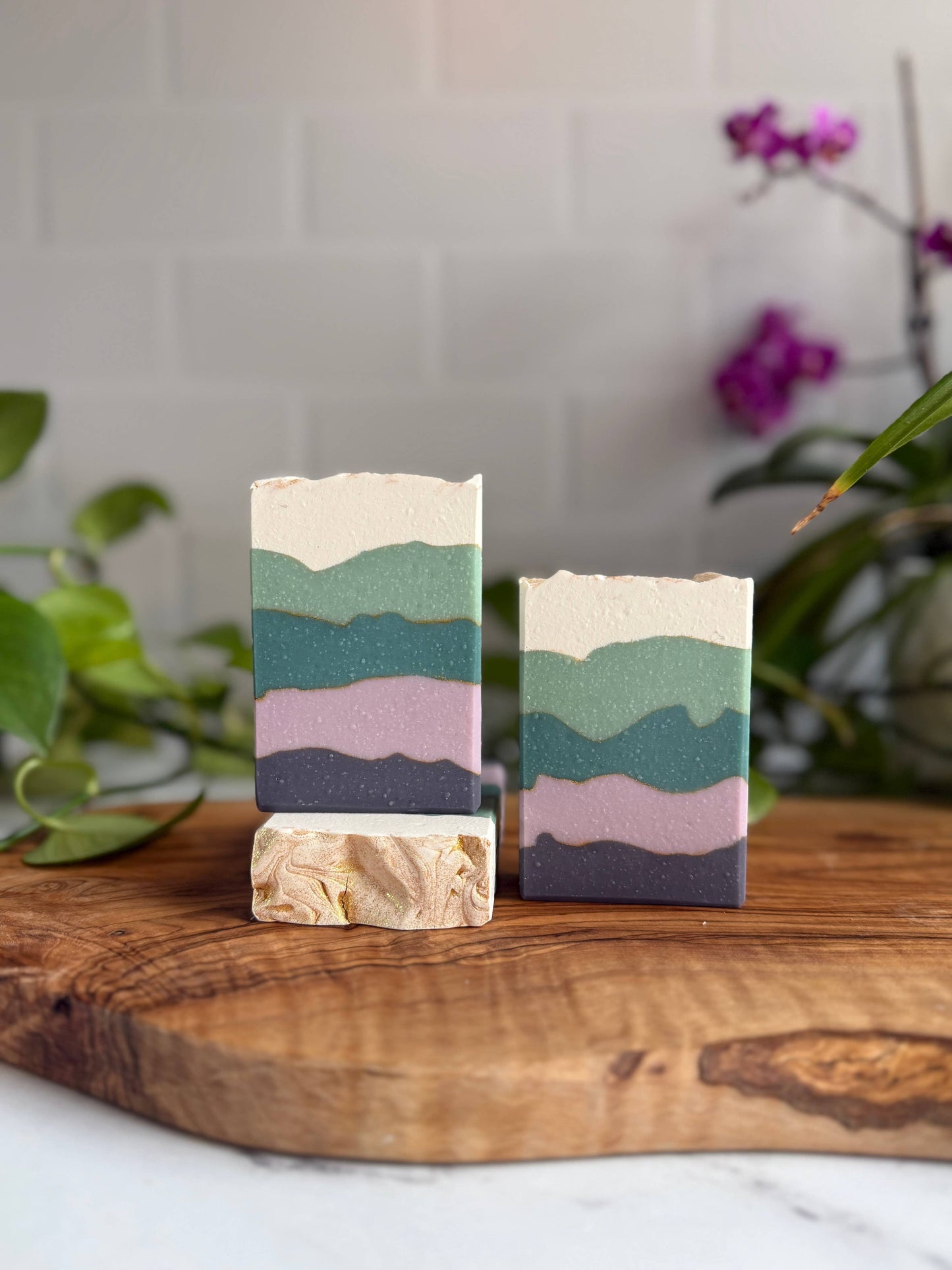 Two blackberry sage soap bars standing up, the left one slightly elevated by standing on a third one laying down. Each soap bar has five irregularly shaped bands, colored top to bottom: cream, light teal, dark teal, light purple, dark purple.