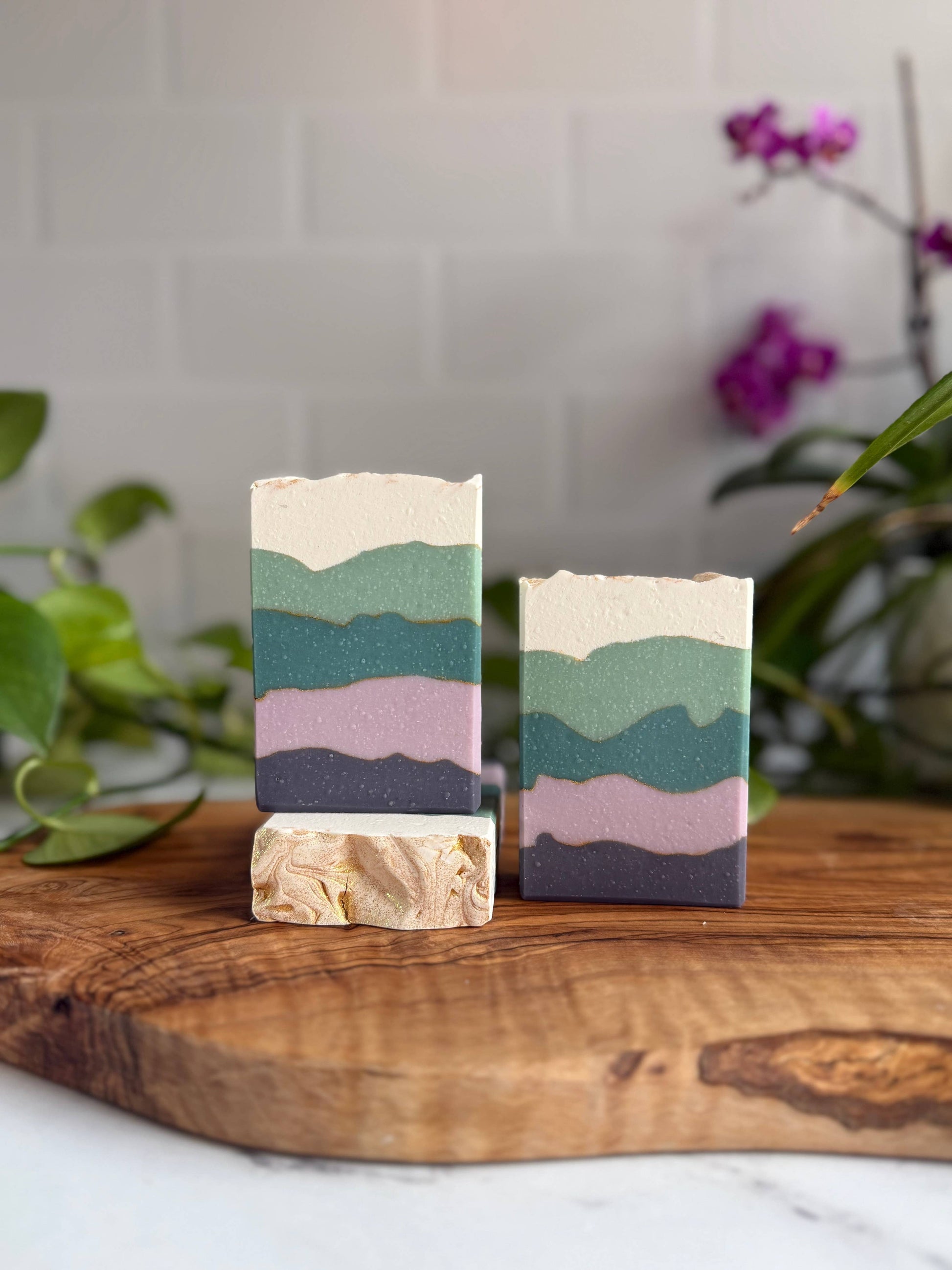 Two blackberry sage soap bars standing up, the left one slightly elevated by standing on a third one laying down. Each soap bar has five irregularly shaped bands, colored top to bottom: cream, light teal, dark teal, light purple, dark purple.