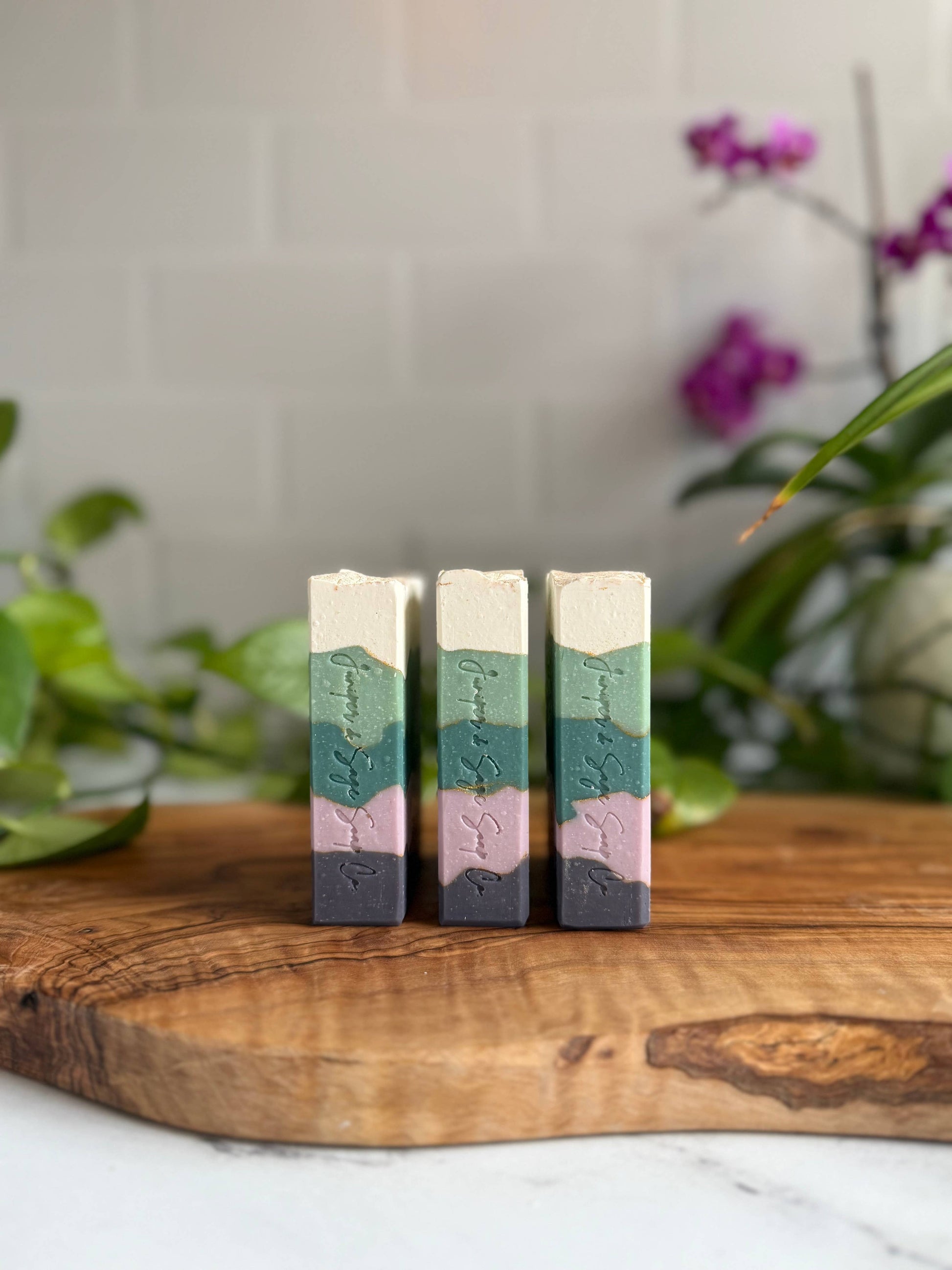 Three blackberry sage soap bars standing up, viewed in side profile. Each soap bar has five irregularly shaped bands, colored top to bottom: cream, light teal, dark teal, light purple, dark purple. The brand name “Juniper & Sage Co” is written on the side.