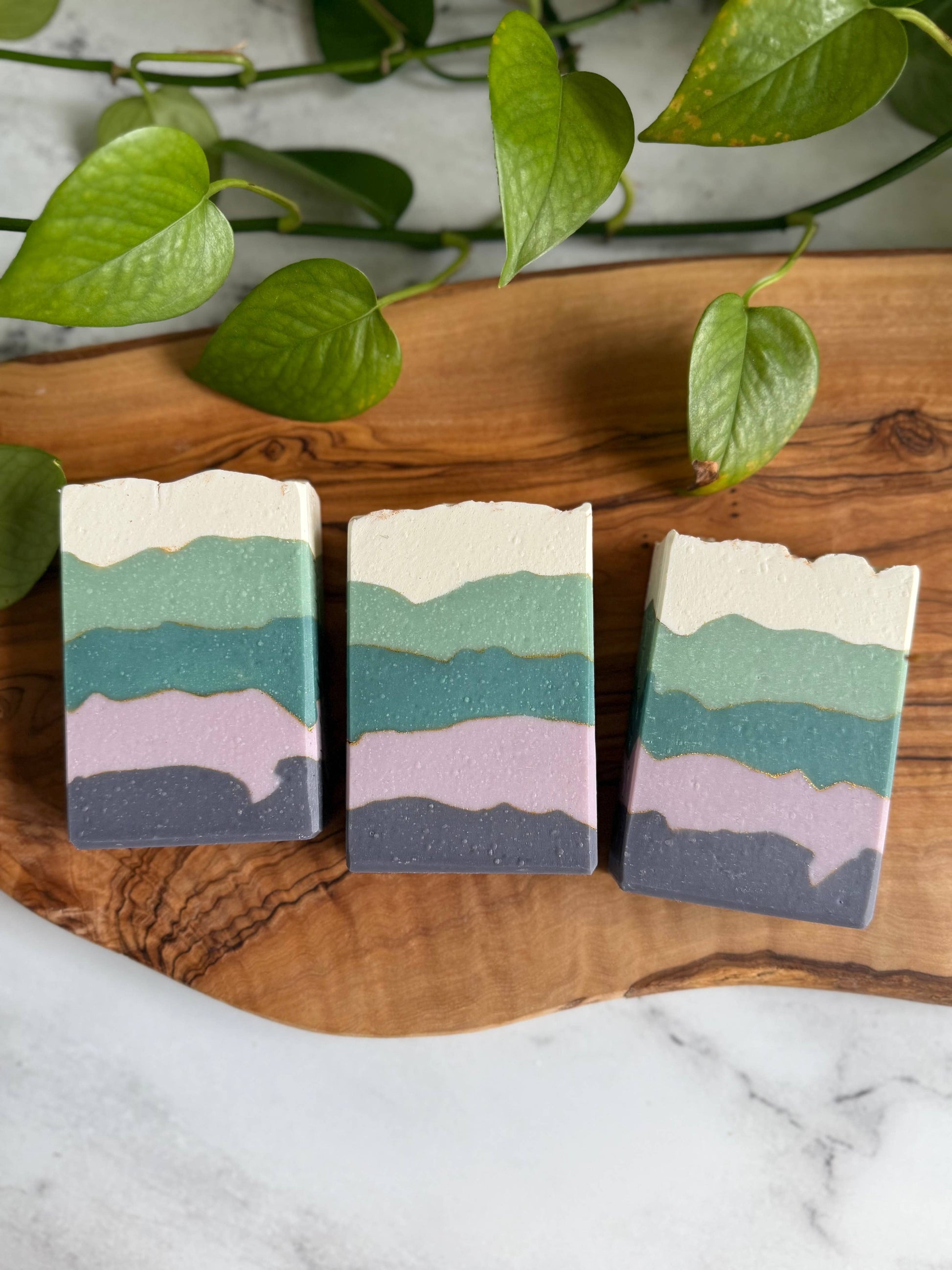 Three blackberry sage soap bars laying down, viewed from above. Each soap bar has five irregularly shaped bands, colored top to bottom: cream, light teal, dark teal, light purple, dark purple.