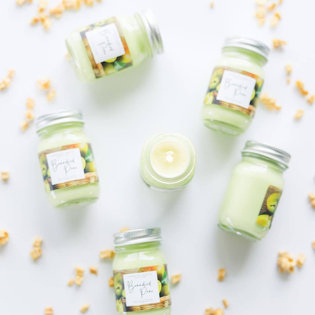 Image of six Brandied Pear candles, pastel  green candles in miniature mason jars. One is open and lit, while the other five are arrayed around it.