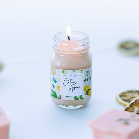 Image of the Citrus Agave candle, a pale pink candle in a miniature mason jar. The jar is open and the candle is lit.