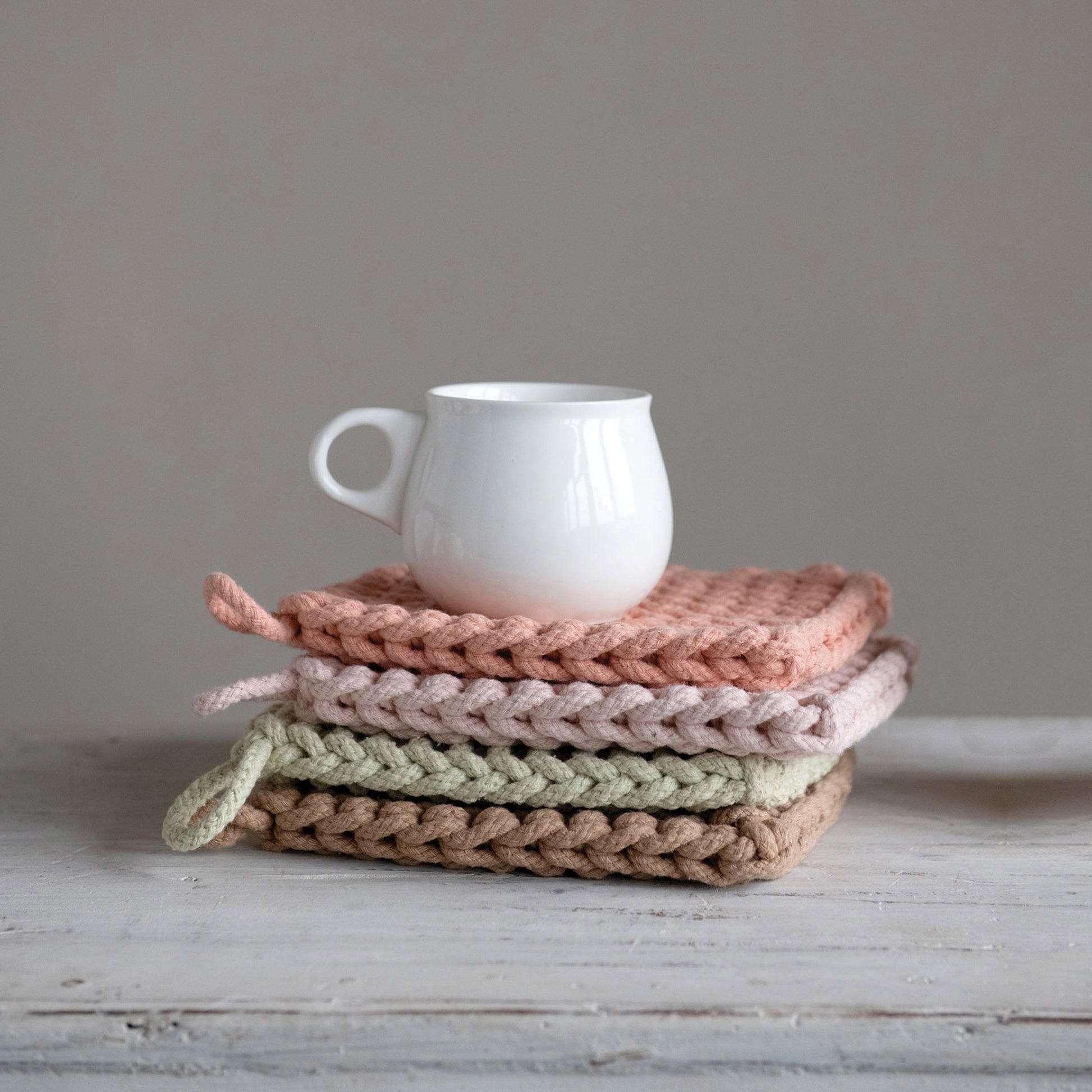 Four crocheted pot holders stacked on top of each other, with a white porcelain cup on top of them. From top to bottom, the pot holders are dark pink, light pink, green, and beige.