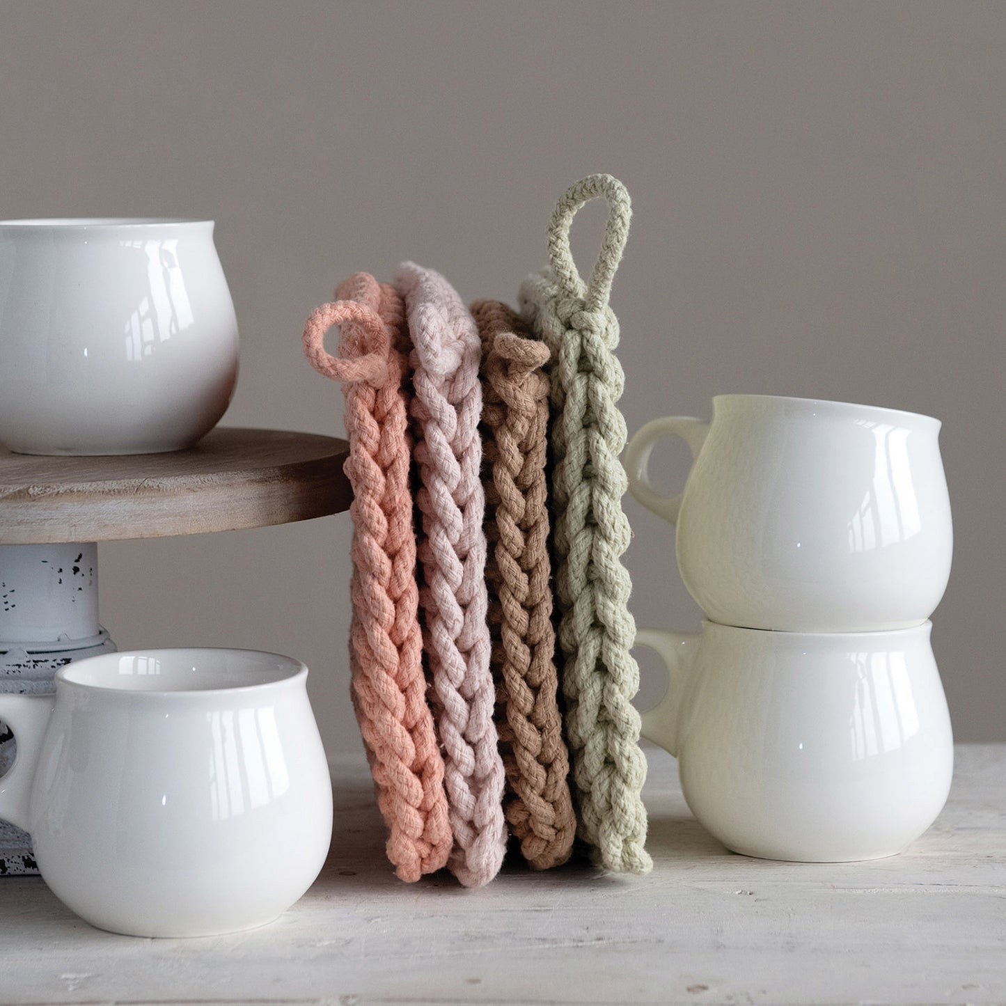 Four crocheted pot holders, lined up perpendicular to the table, leaning against a cake stand, surrounded by porcelain cups. From left to right, the pot holders are dark pink, light pink, beige, and green.