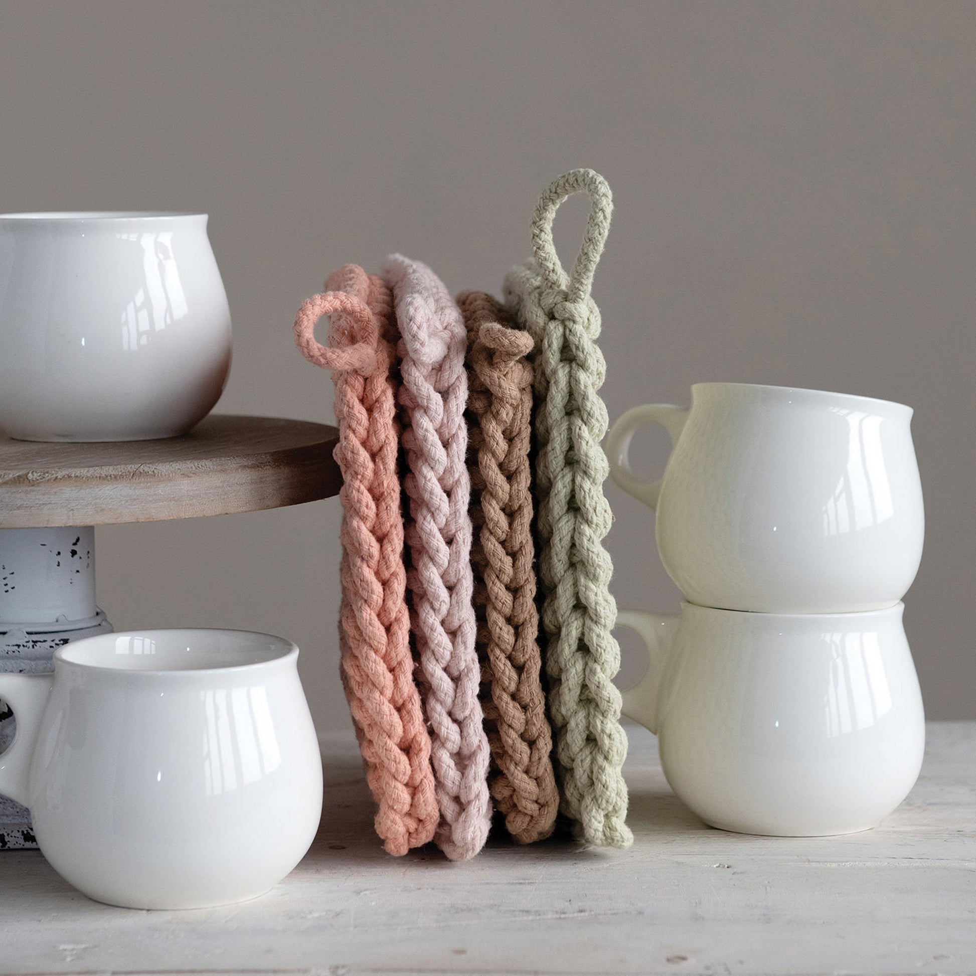 Four crocheted pot holders, lined up perpendicular to the table, leaning against a cake stand, surrounded by porcelain cups. From left to right, the pot holders are dark pink, light pink, beige, and green.