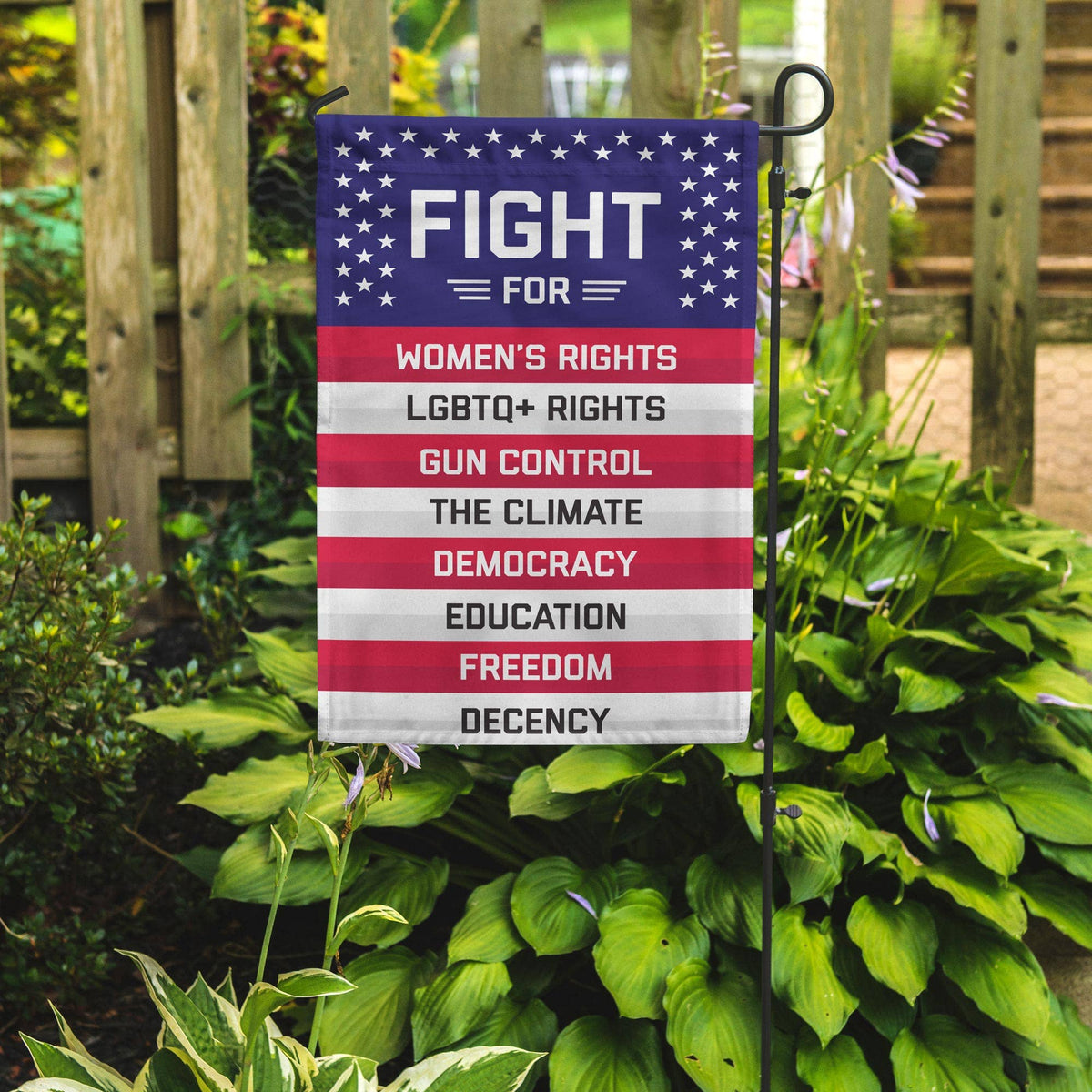 A lawn flag styled after the USA flag, with the text “FIGHT FOR WOMEN’S RIGHTS, LGBTQ+ RIGHTS, GUN CONTROL, THE CLIMATE, DEMOCRACY, EDUCATION, FREEDOM, DECENCY”. The flag is displayed in front of some greenery and a patio.