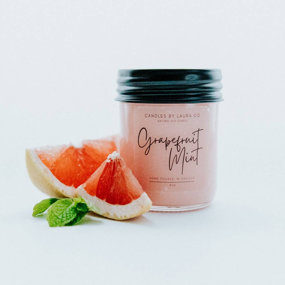 A pastel pink candle in a glass jar labeled “Grapefruit Mint”, alongside a slice of grapefruit and a sprig of mint.