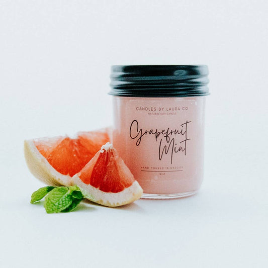 A pastel pink candle in a glass jar labeled “Grapefruit Mint”, alongside a slice of grapefruit and a sprig of mint.
