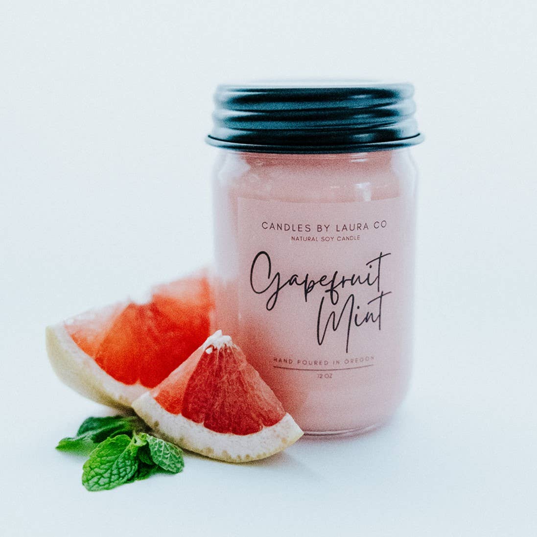 A pastel pink candle in a glass jar labeled “Grapefruit Mint”, alongside a slice of grapefruit and a sprig of mint.