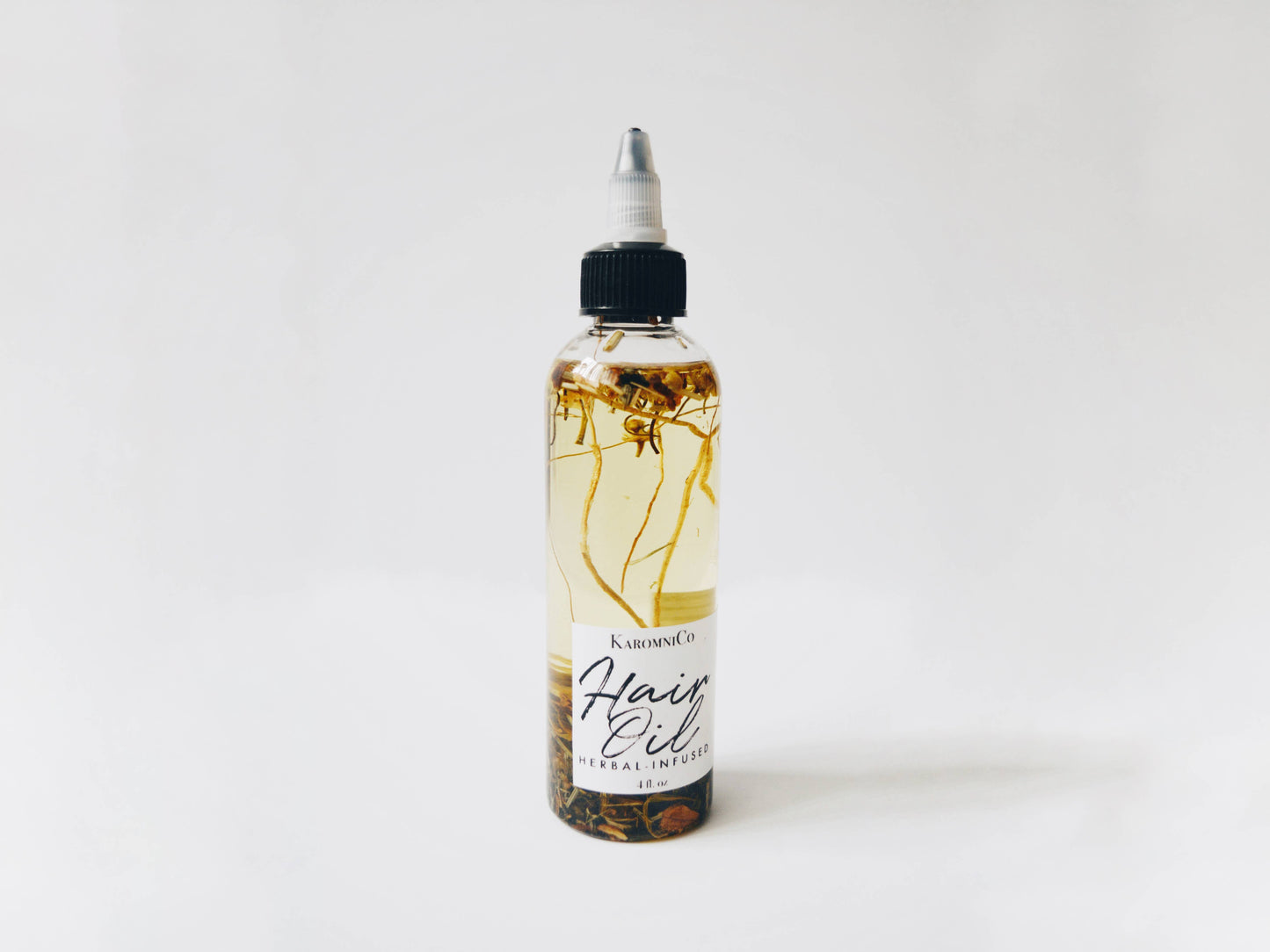 Shows the hair oil against a white background. It’s a yellowish oil with various small herbs and fragments of plants floating in it.