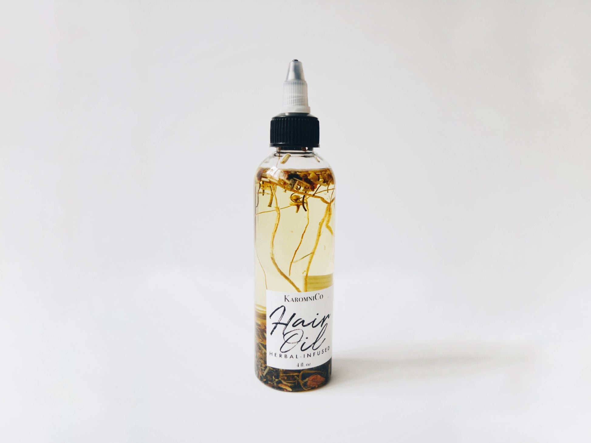Shows the hair oil against a white background. It’s a yellowish oil with various small herbs and fragments of plants floating in it.