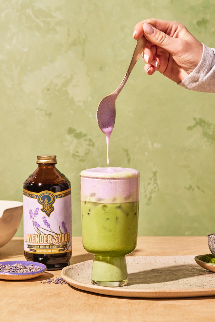 A bottle of lavender syrup next to a glass filled with a matcha drink that is topped with a purple foam. A person’s hand holds a spoon over the drink, which has had some of the foam scooped onto it.