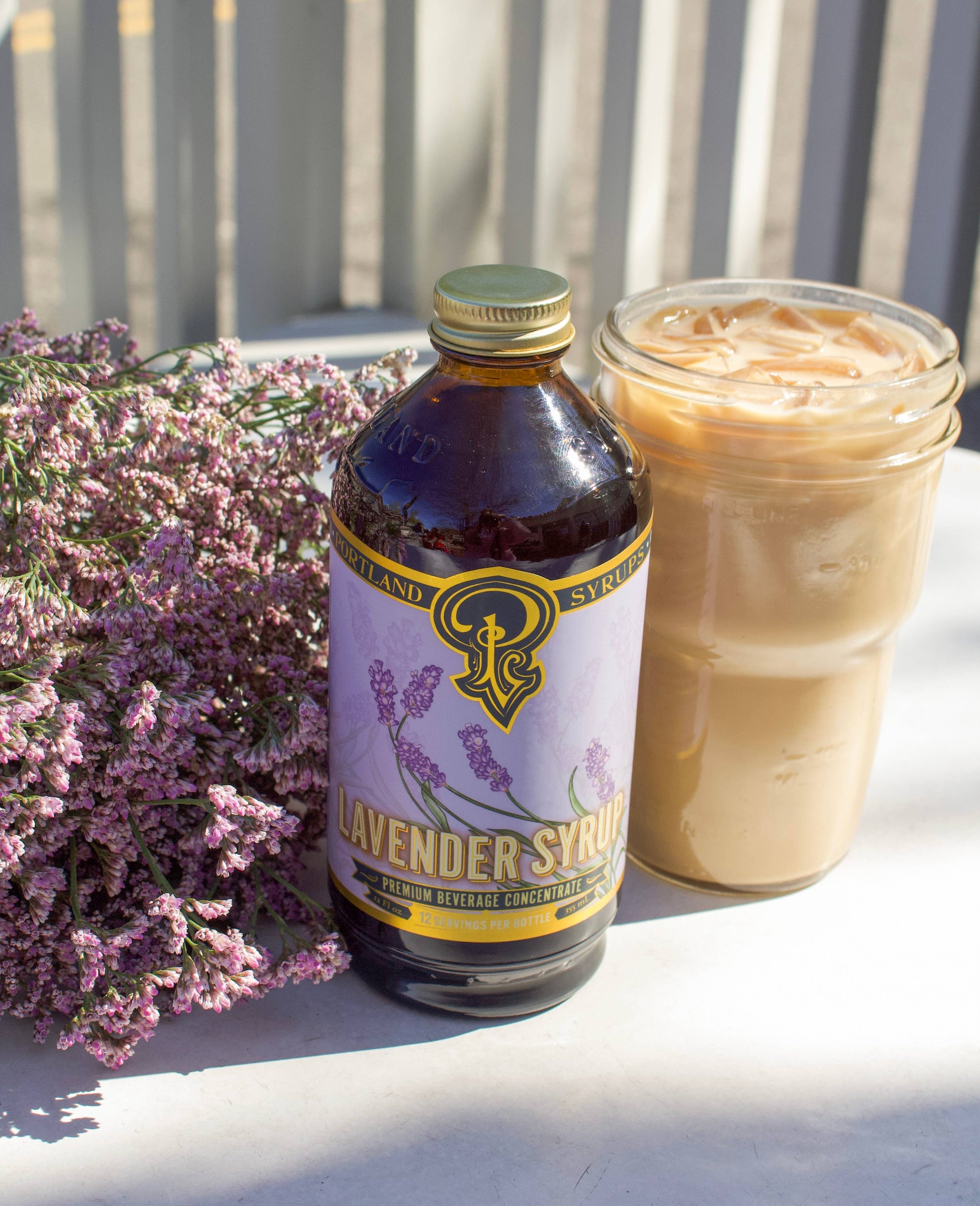 A dark glass bottle labeled “Portland Syrups - Lavender Syrup - Premium Beverage Concentrate”. It sits out in the sunlight next to a glass of iced coffee and a bundle of lavender flowers.