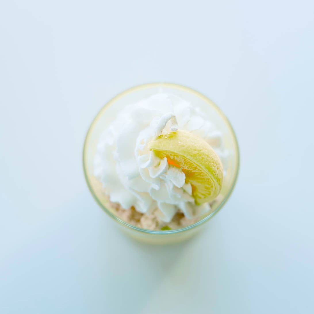 Top-down view of a candle in a clear glass jar, clearly showing that it is topped with decorations of whipped cream and a slice of lemon, both sculpted from wax.
