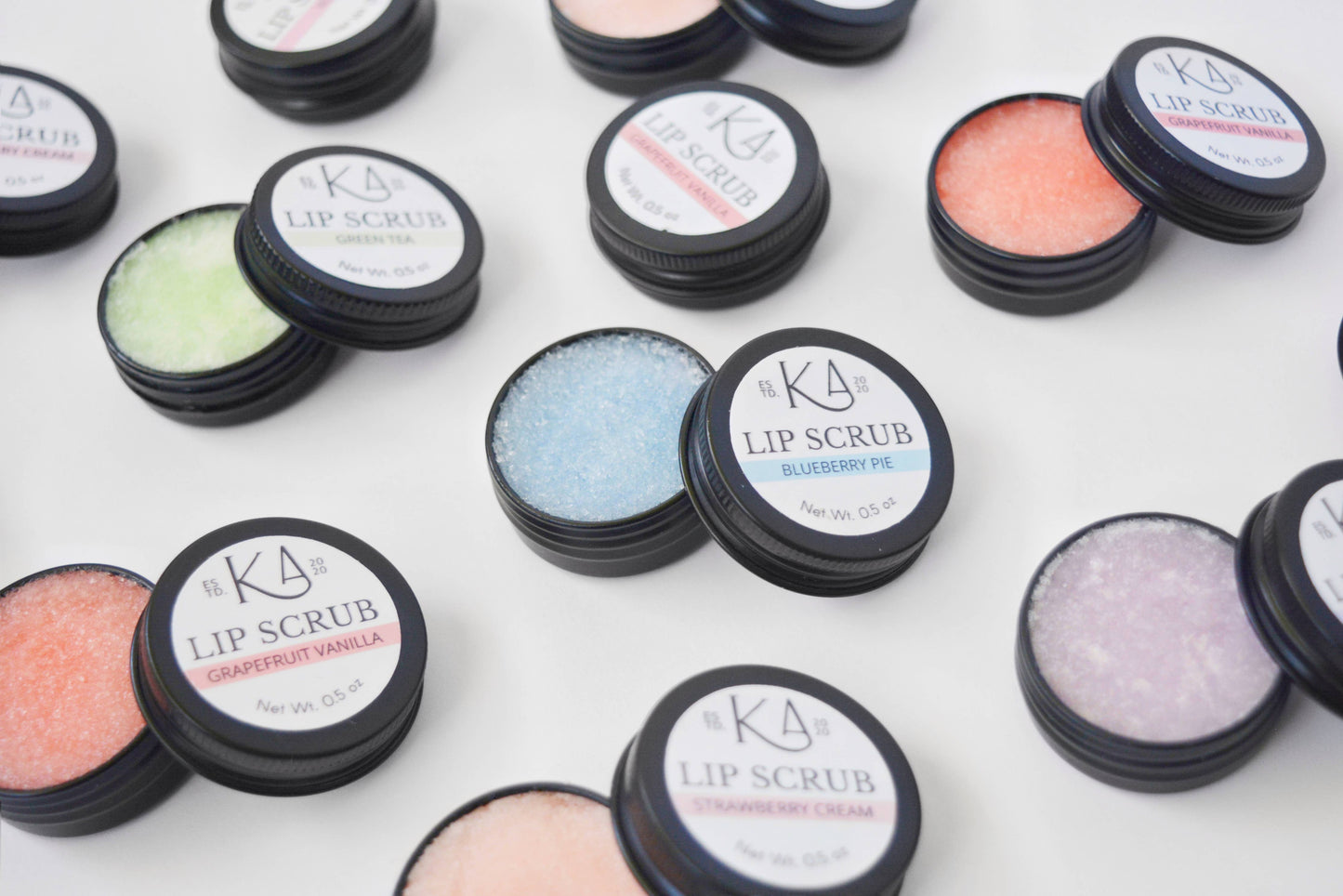 Shows an assortment of lip scrub tins against a white background. They’re cracked open, revealing the scrubs inside, which are a variety of pastel colors.