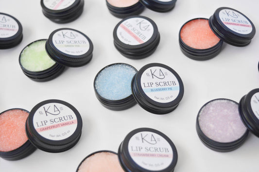 Shows an assortment of lip scrub tins against a white background. They’re cracked open, revealing the scrubs inside, which are a variety of pastel colors.