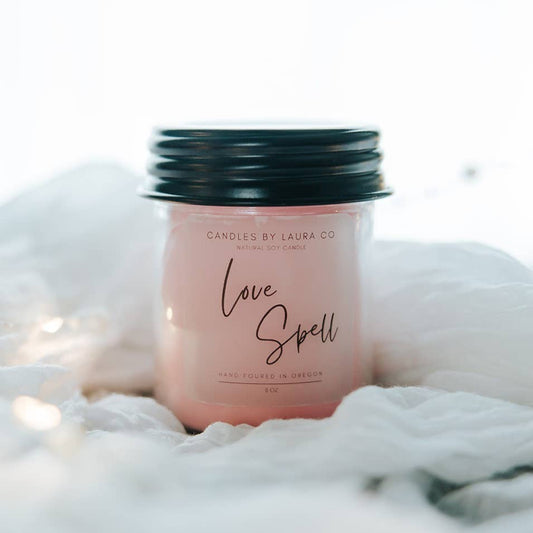 A pink candle in a jar with a black lid, labeled “Love Spell”.