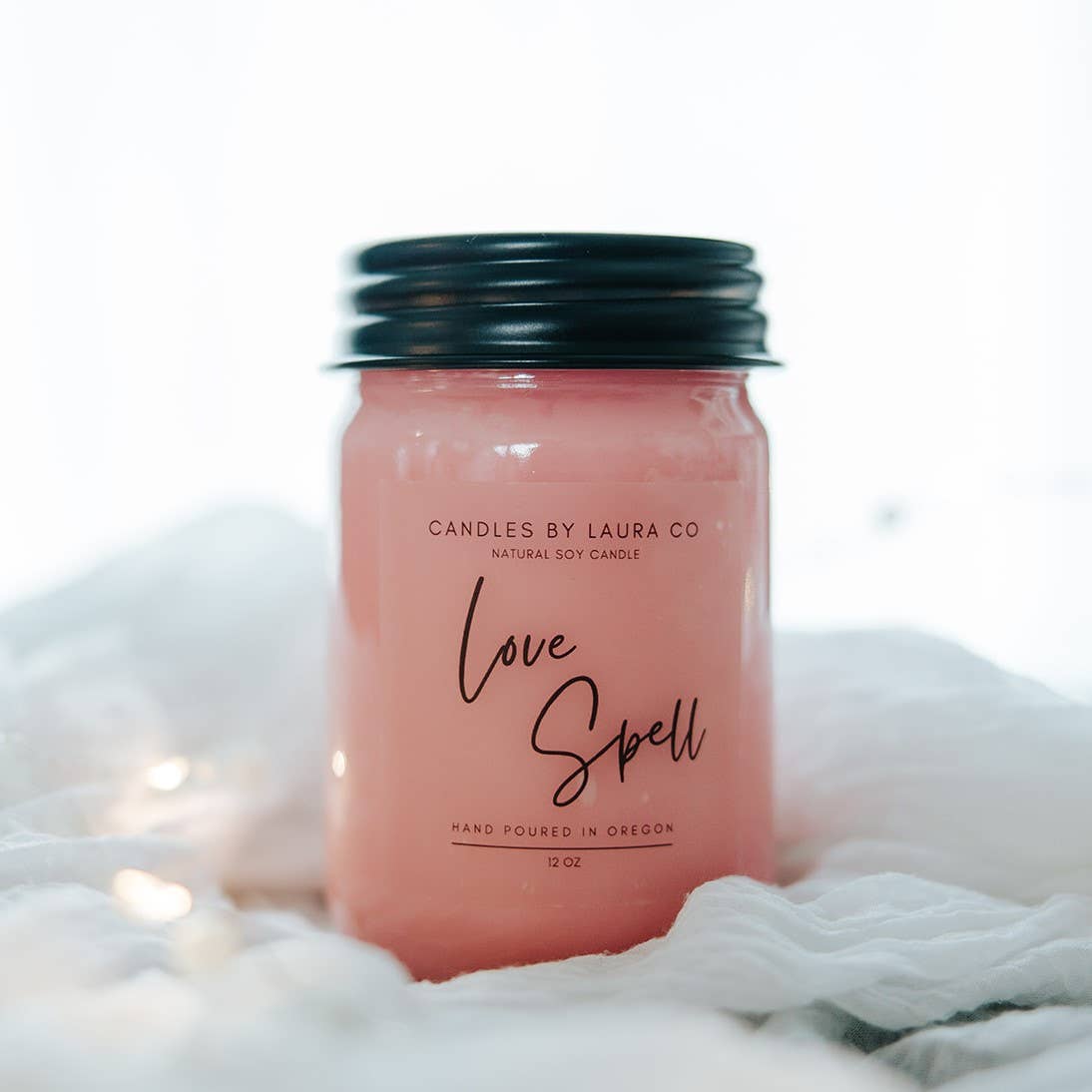A pink candle in a jar with a black lid, labeled “Love Spell”.