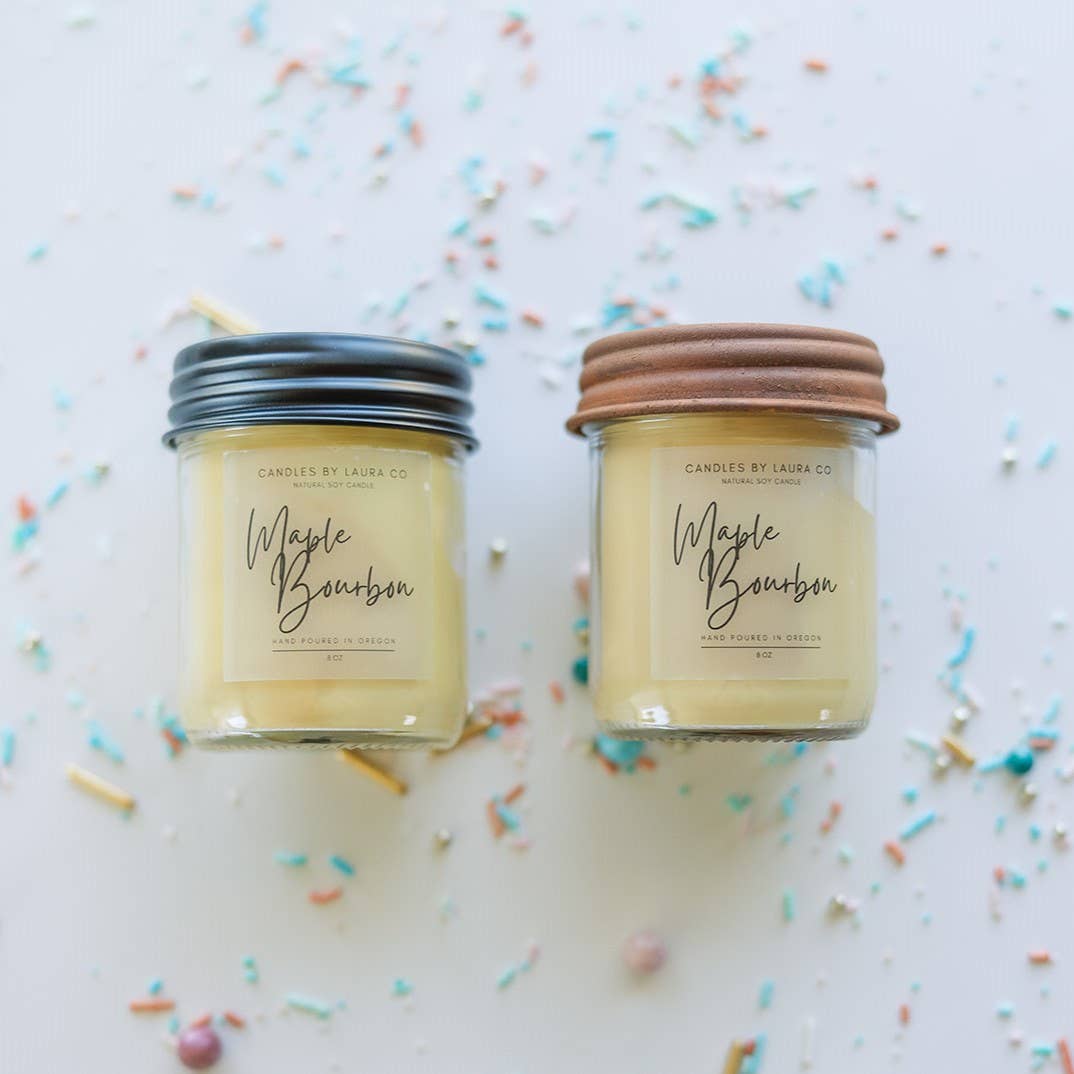 Image of two Maple Bourbon candles, off-white candles in jars; the left candle has a black lid while the right candle has a light brown lid.