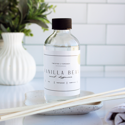 A glass bottle of clear liquid with two thin reeds laying in front of it. The bottle is labeled “Vanilla Bean Reed Diffuser”.