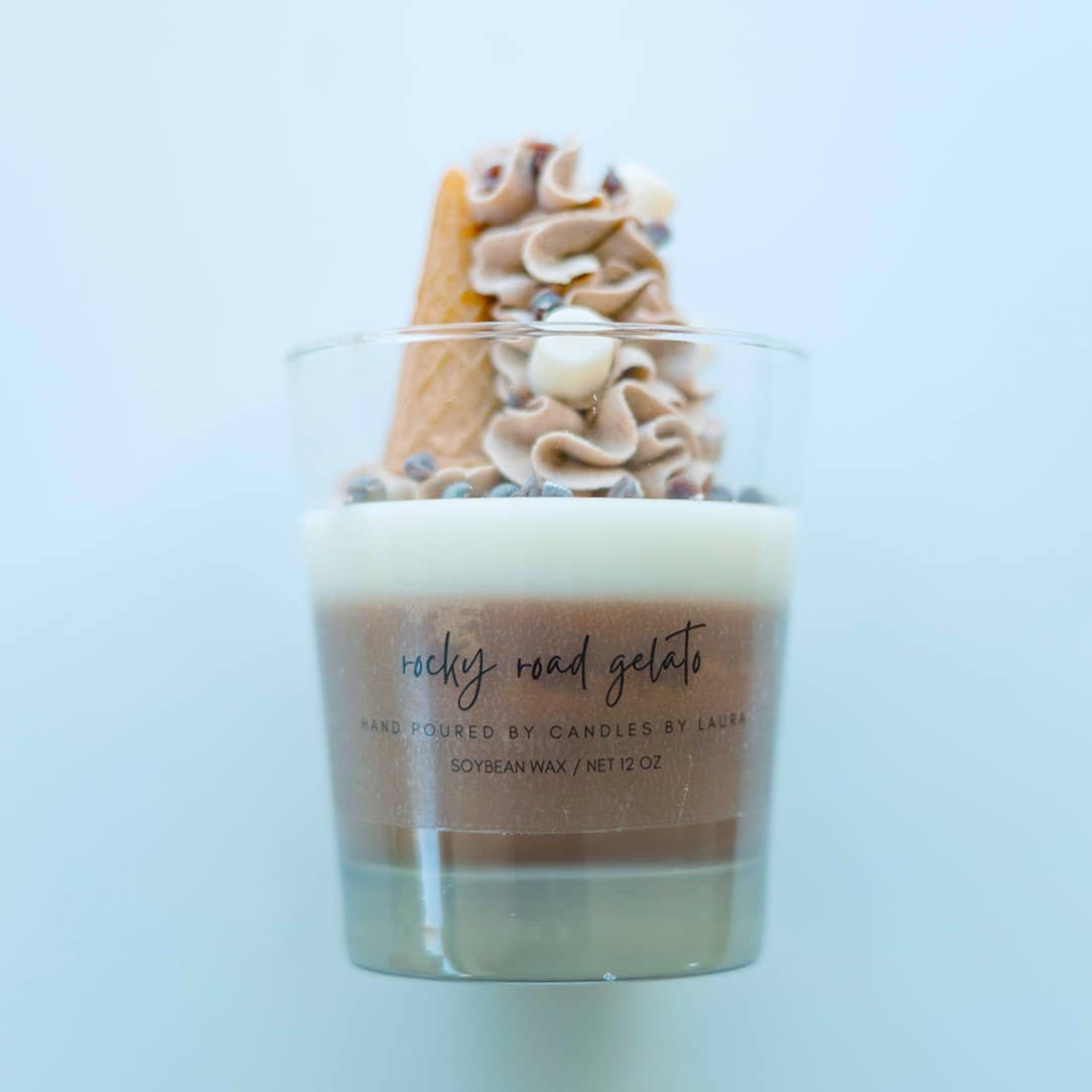 A candle in a clear glass jar. The bottom layer of the candle is chocolate brown, and the top layer is white. It’s topped with decorations of marshmallows, chocolate chips, chocolate-colored whipped cream, and an upside-down ice cream cone, all sculpted from wax.
