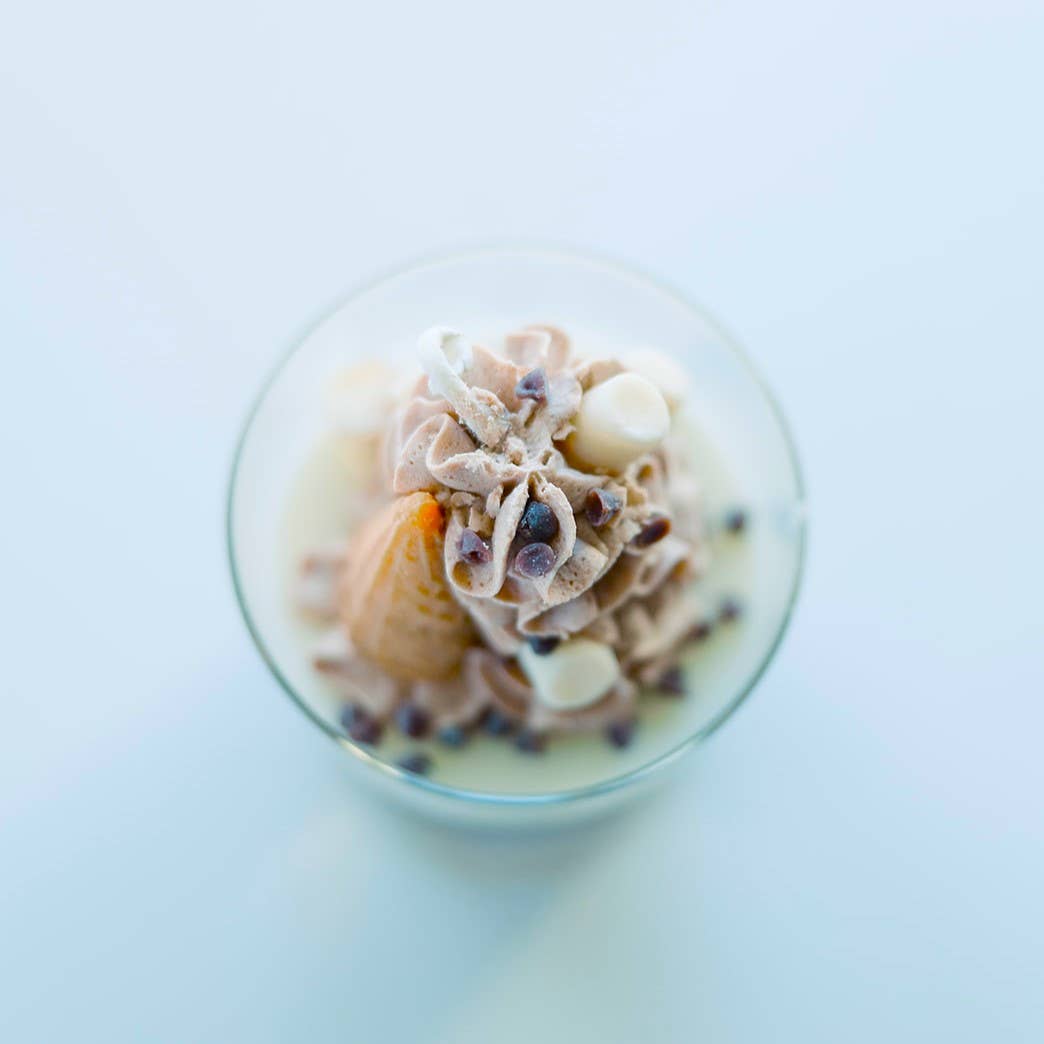 A top-down view of a candle in a clear glass jar, clearly showing that it is topped with decorations of marshmallows, chocolate chips, chocolate-colored whipped cream, and an upside-down ice cream cone, all sculpted from wax.