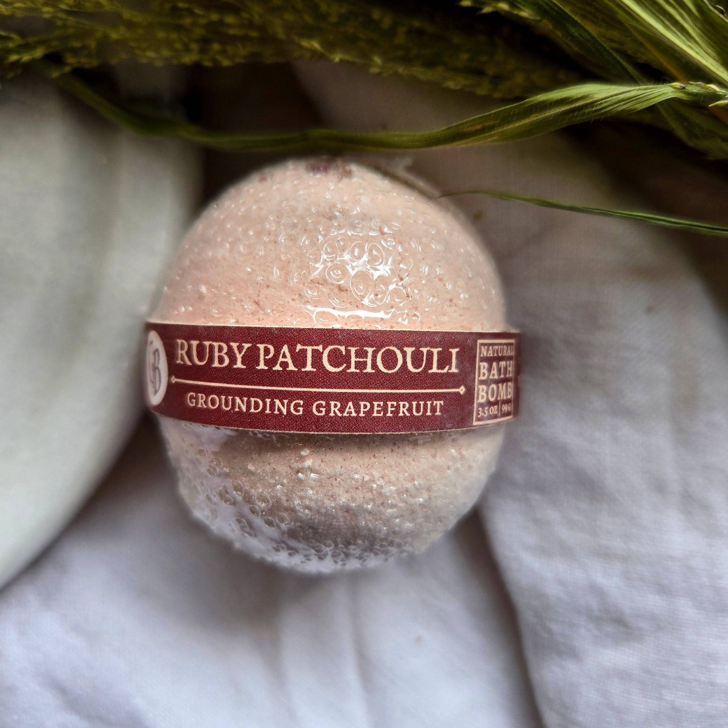 A pale pink bath bomb labeled as “Ruby Patchouli”, with the tagline “Grounding Grapefruit.”