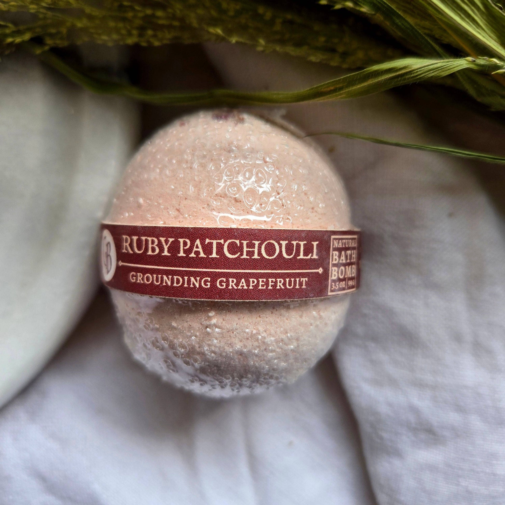 A pale pink bath bomb labeled as “Ruby Patchouli”, with the tagline “Grounding Grapefruit.”