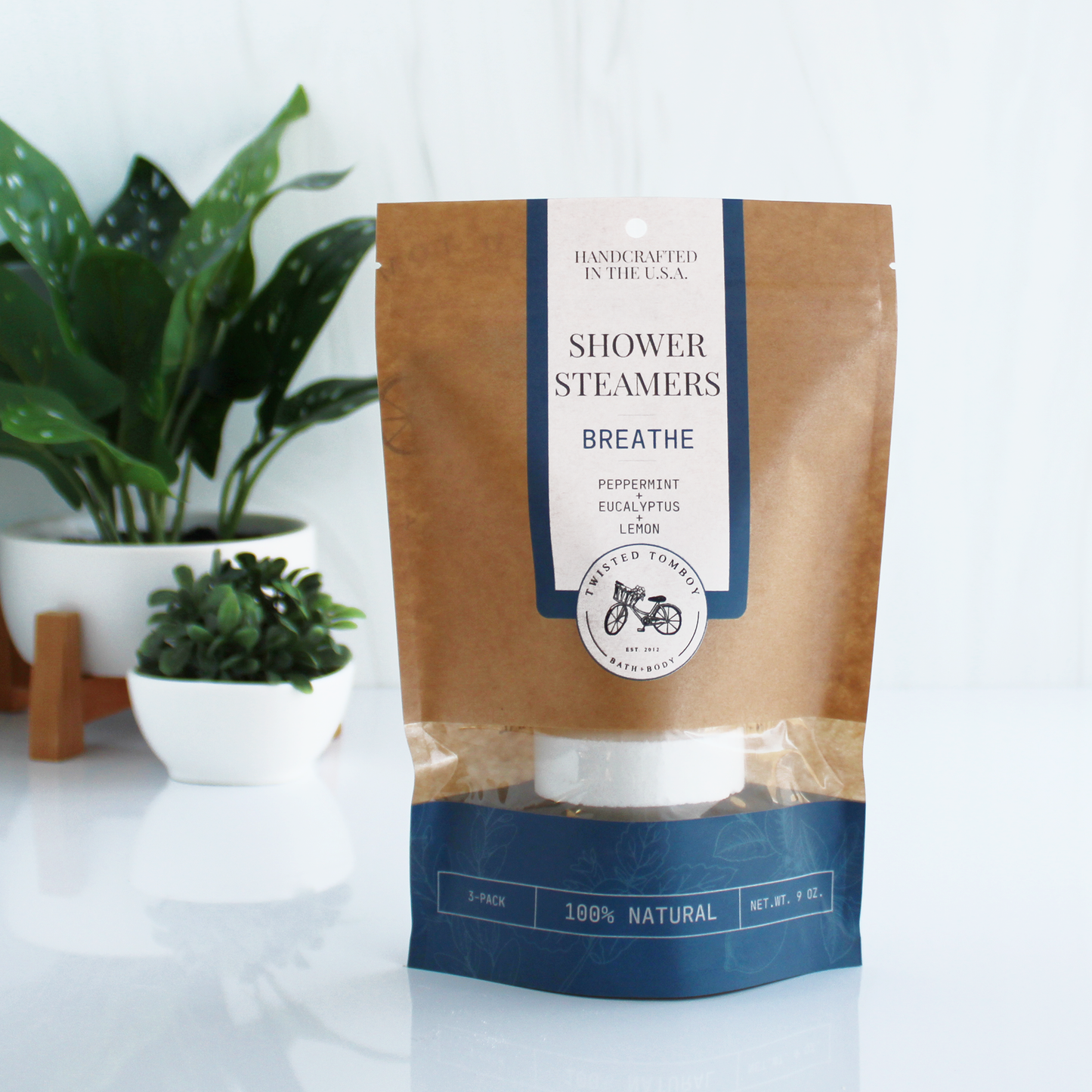 A brown bag with blue accents labeled “Shower Steamers - Breathe - Peppermint+Eucalyptus+Lemon”. Part of the bag is clear, showing that it contains a shower steamer (a thick white tablet).