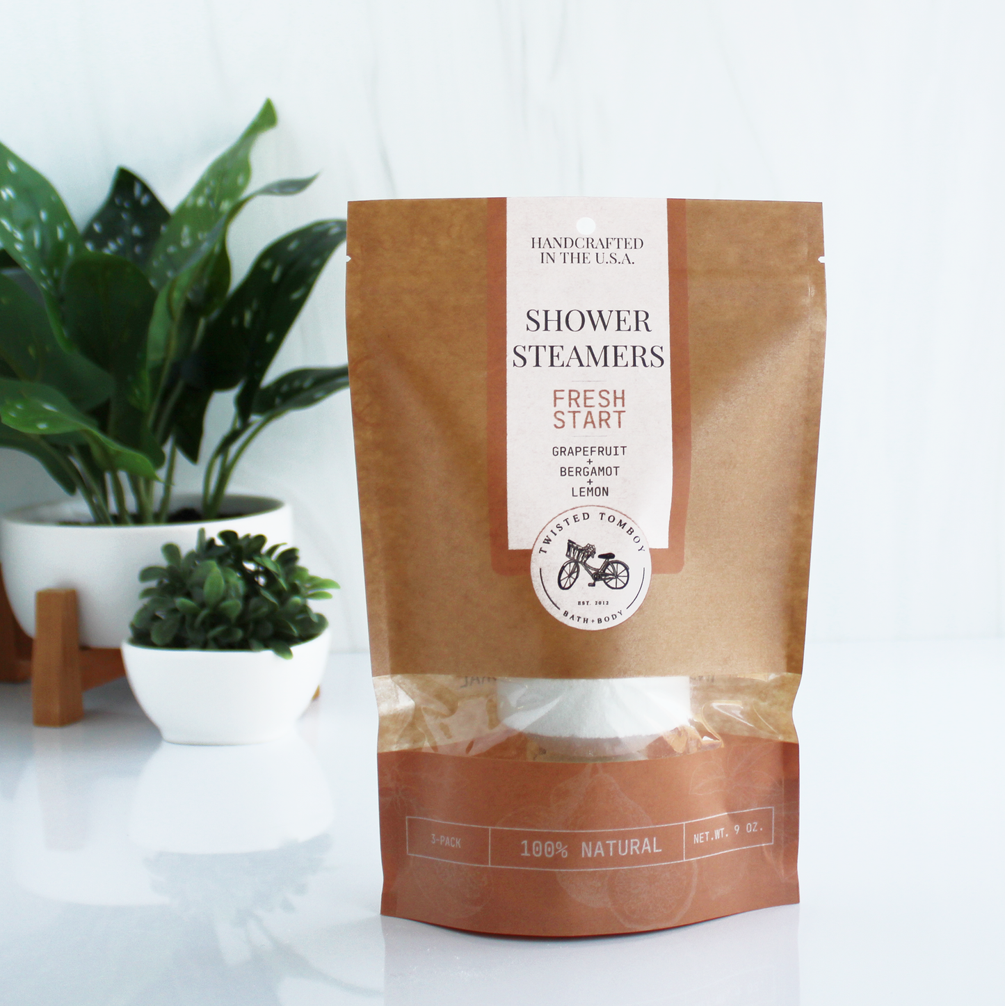 A brown bag with salmon-colored accents labeled “Shower Steamers - Fresh Start - Grapefruit+Bergamot+Lemon”. Part of the bag is clear, showing that it contains a shower steamer (a thick white tablet).