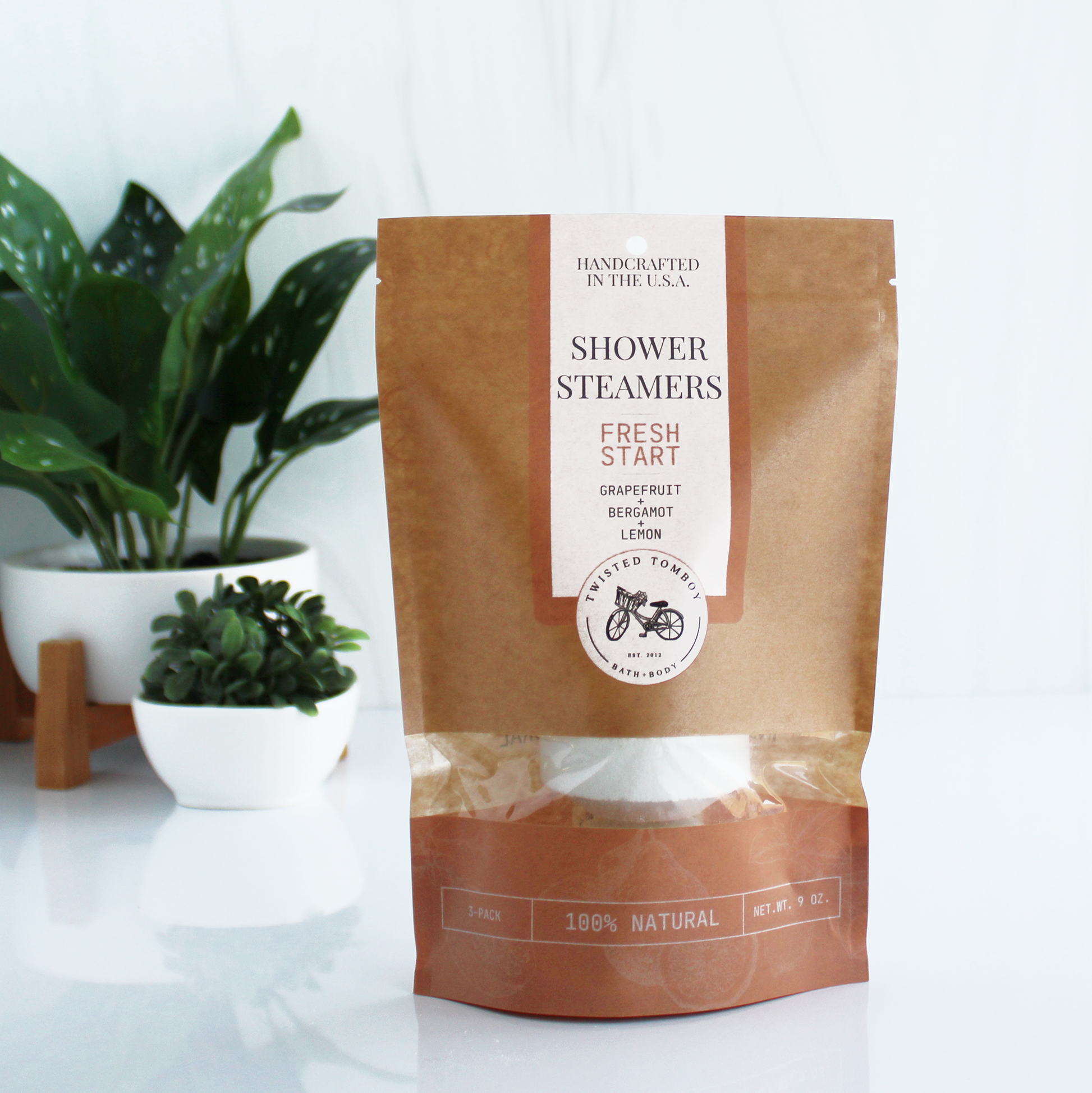 A brown bag with salmon-colored accents labeled “Shower Steamers - Fresh Start - Grapefruit+Bergamot+Lemon”. Part of the bag is clear, showing that it contains a shower steamer (a thick white tablet).