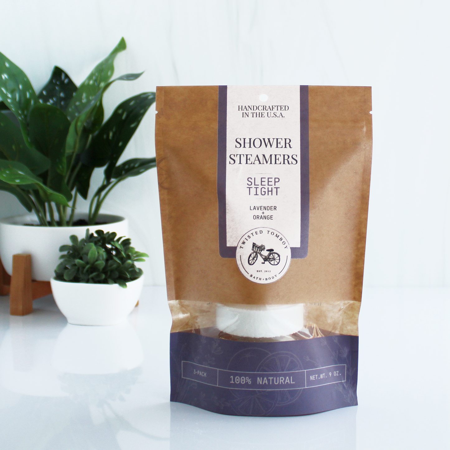 A brown bag with purple accents labeled “Shower Steamers - Sleep Tight - Lavender+Orange”. Part of the bag is clear, showing that it contains a shower steamer (a thick white tablet).