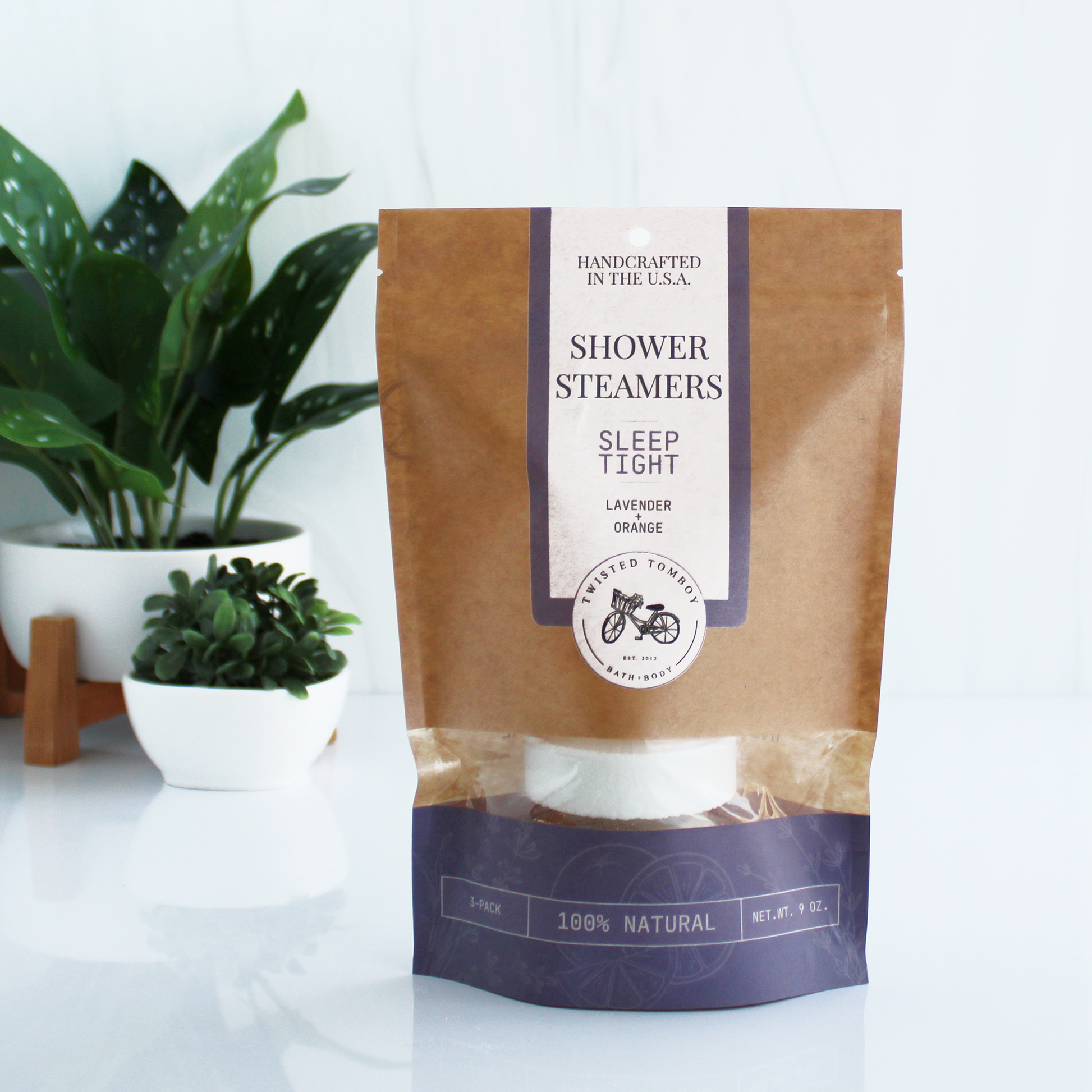 A brown bag with purple accents labeled “Shower Steamers - Sleep Tight - Lavender+Orange”. Part of the bag is clear, showing that it contains a shower steamer (a thick white tablet).