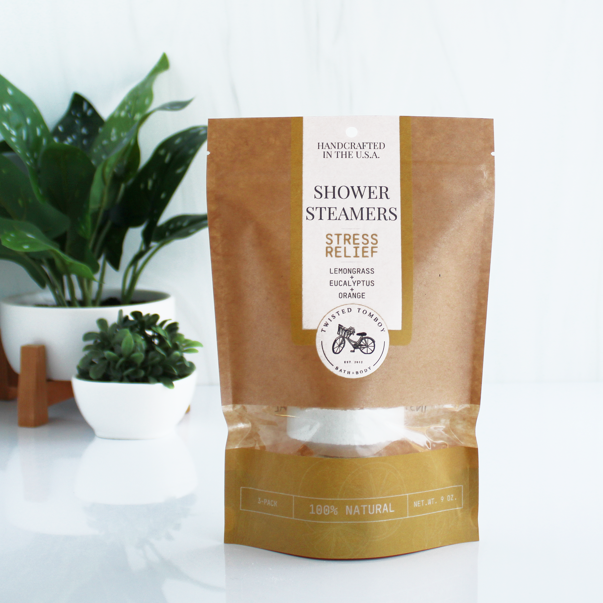 A brown bag with yellow accents labeled “Shower Steamers - Stress Relief - Lemongrass+Eucalyptus+Orange”. Part of the bag is clear, showing that it contains a shower steamer (a thick white tablet).