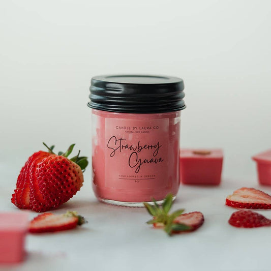 A pinkish red candle in a glass jar labeled “Strawberry Guava” surrounded by slices of strawberry.
