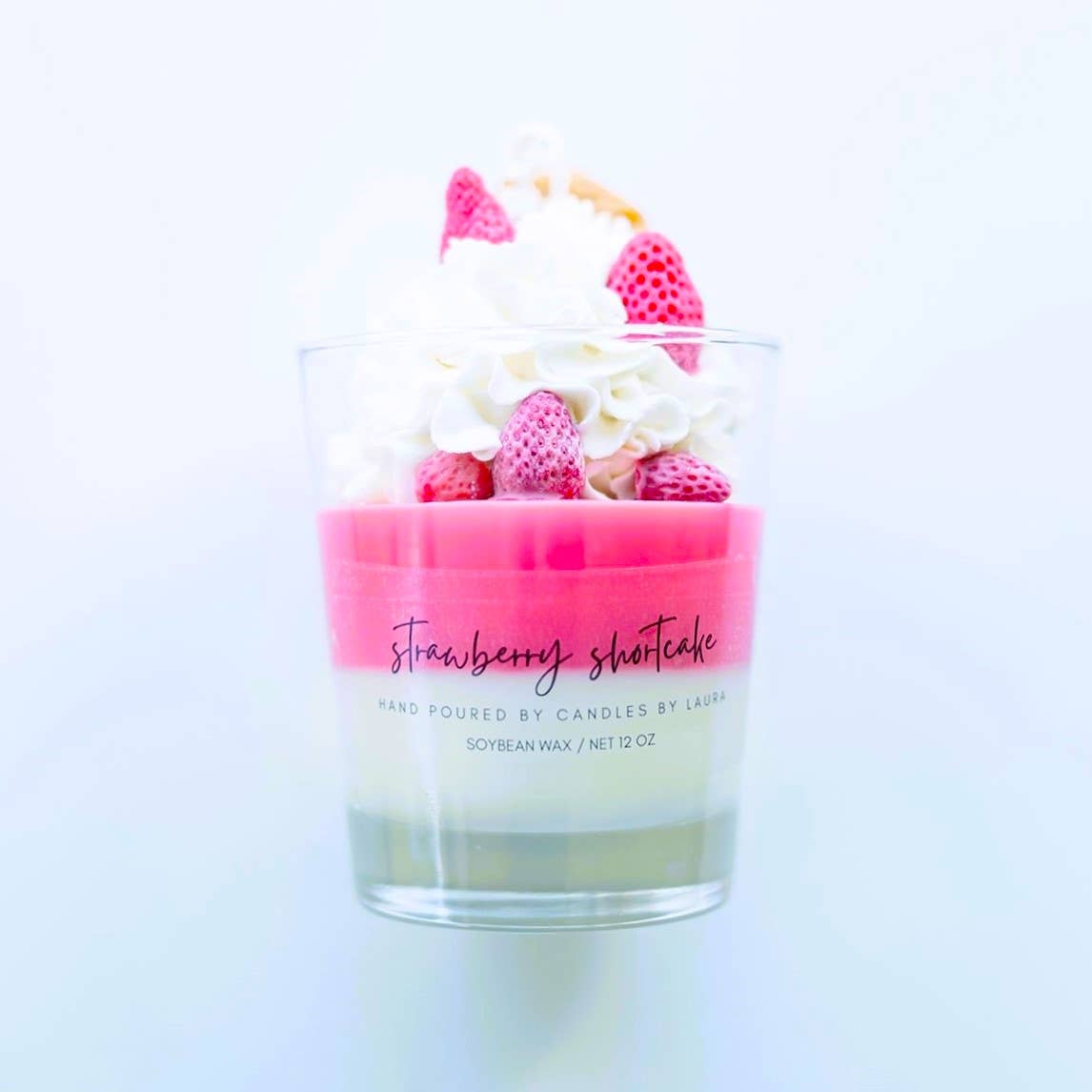 Image of a candle in a clear glass jar. The bottom layer of it is white wax and the top layer is hot pink wax. On top of those layers is decorations of whipped cream, strawberry, and wafers, all sculpted from wax.