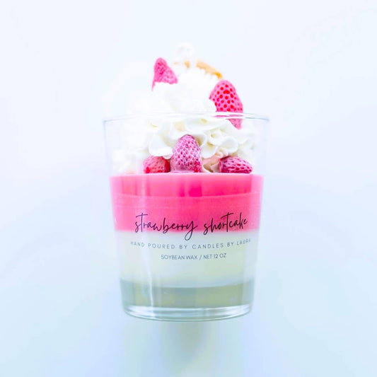 Image of a candle in a clear glass jar. The bottom layer of it is white wax and the top layer is hot pink wax. On top of those layers is decorations of whipped cream, strawberry, and wafers, all sculpted from wax.