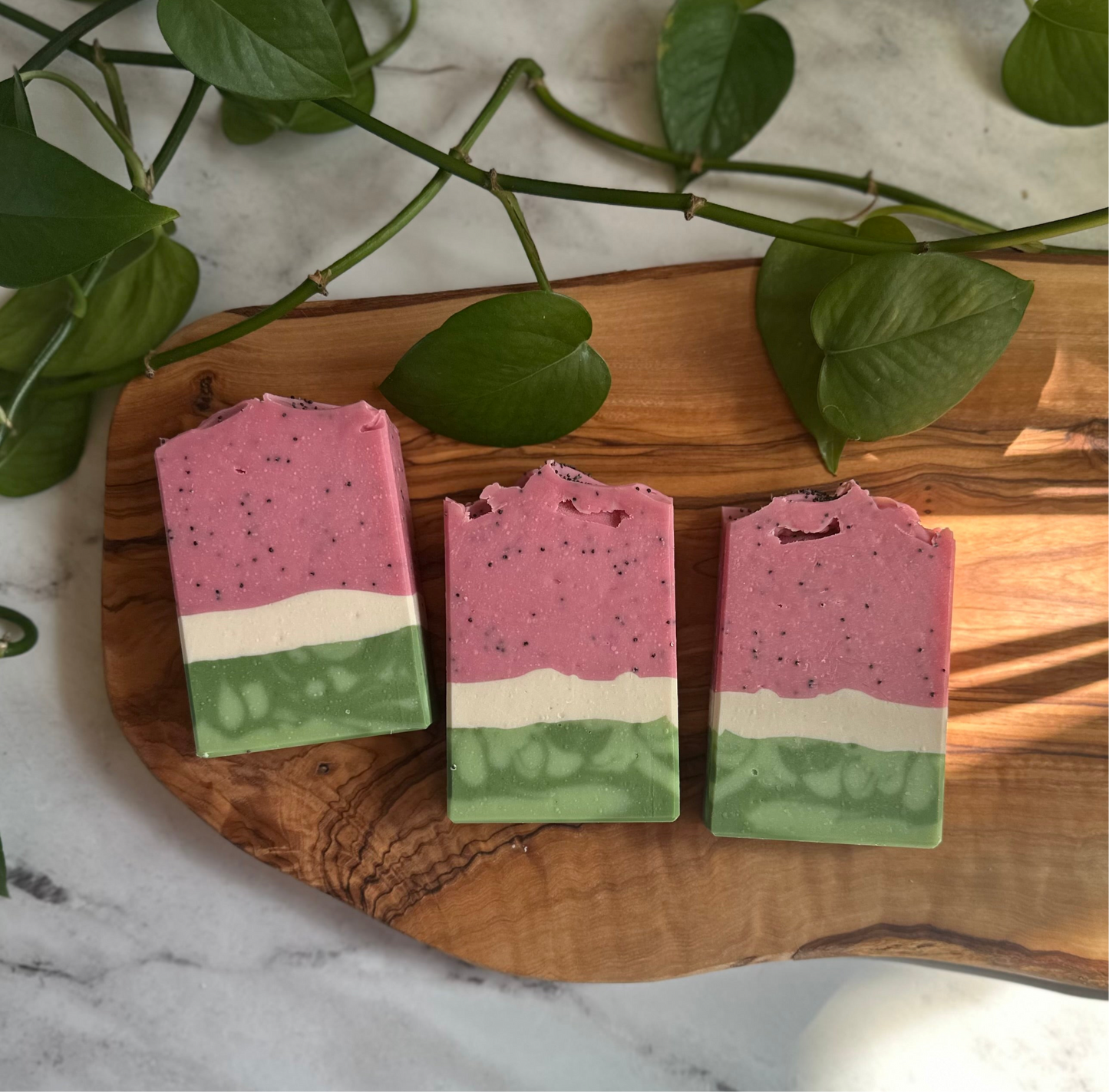 Three watermelon soap bars laying down, viewed from above, side-by-side with each other. Each soap bar resembles a slice of watermelon, with a thick band of pink speckled with poppy seeds, a thin band of white, and a slightly thicker band of green.