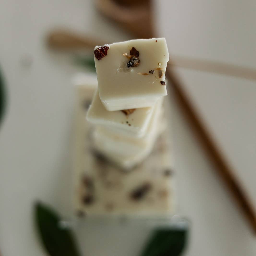 Image of the White Oak & Vanilla wax melt, broken apart into six blocks, stacked on top of each other, resting on top of an intact wax melt. The White Oak & Vanilla wax melt is cream-colored wax with pieces of oak bark and vanilla bean suspended in it.