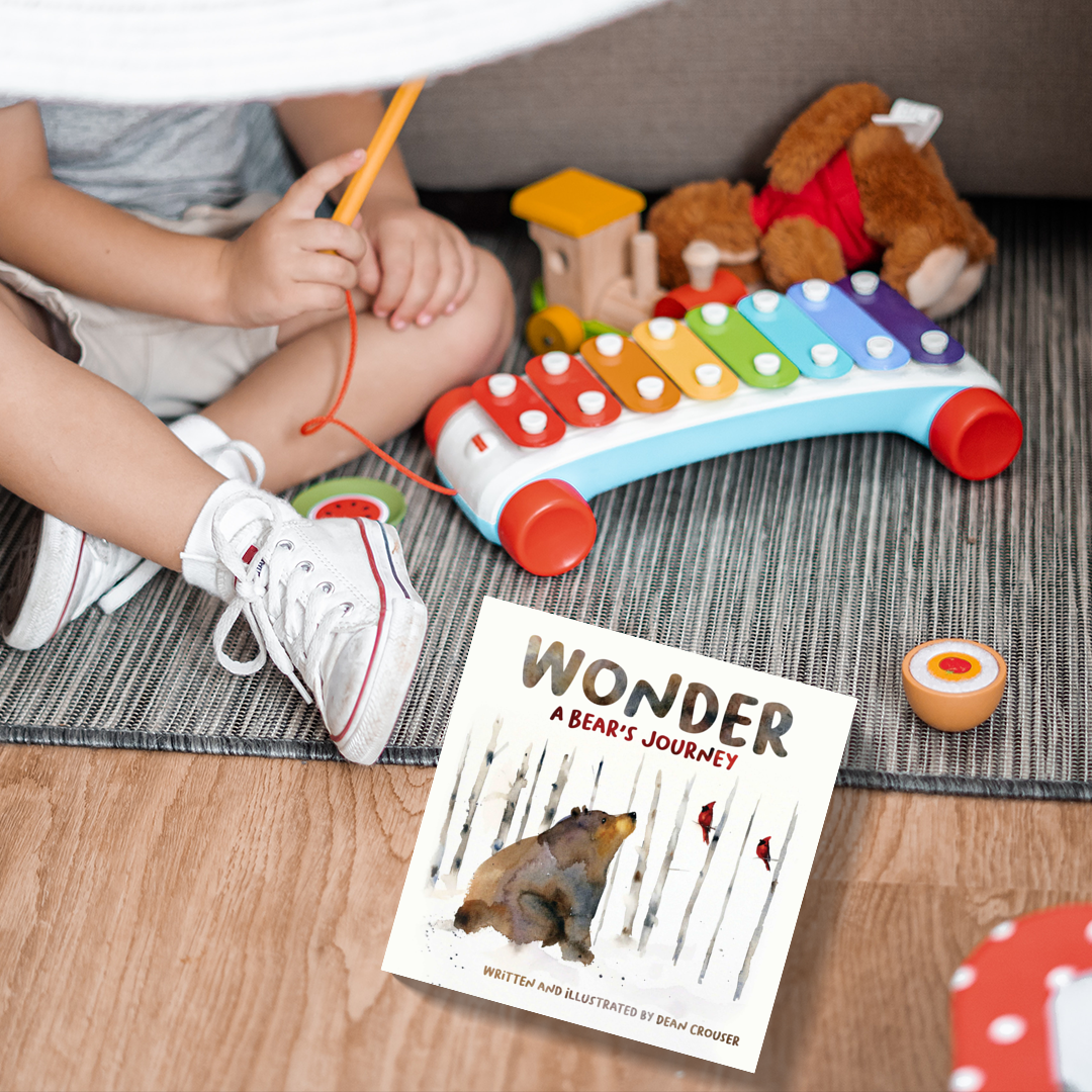 A young child sitting cross-legged on the floors, next to some toys and a copy of the book WONDER: A Bear’s Journey.