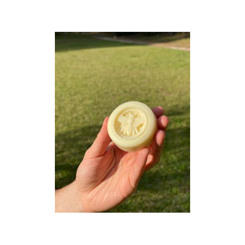 A hand holding a round, pale yellow lotion bar outdoors against a blurred green grass background.