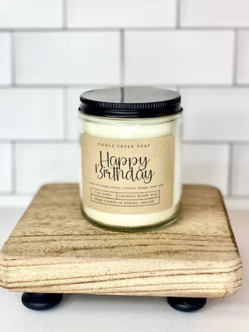Candle jar labeled 'Happy Birthday' on a wooden block with a tiled wall background