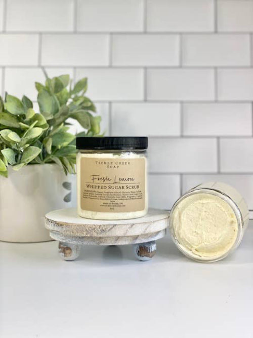 A jar of Lemon Whipped Sugar Scrub placed on a white surface with a white tiled wall in the background. A green plant is also visible to the left.