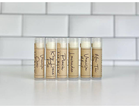 Six lip balm tubes standing upright on a white surface against a white subway tile wall. Each tube has a beige paper label with handwritten-style black text naming different scents: Vanilla Cinnamon, Key Lime, Peach, Lavender, Vanilla Mint, and White Tea.