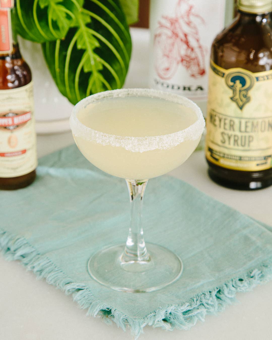 Cocktail in a glass with sugar rim on a teal napkin, surrounded by bottles of cocktail ingredients.