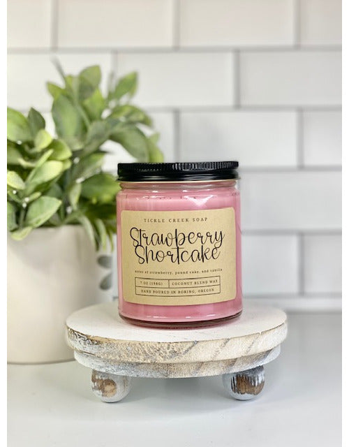 A pink candle in a glass jar with a black lid and a beige label reading 'Strawberry Shortcake' by Tickle Creek Soap, displayed on a round wooden stand against a white subway tile background with a green plant.