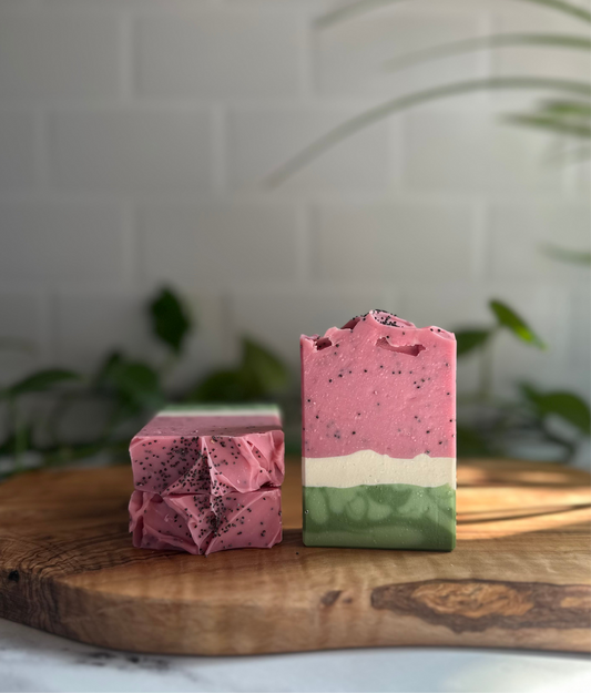 Three watermelon soap bars, with two on the left laying down on their face and one on the right standing up. Each soap bar resembles a slice of watermelon, with a thick band of pink speckled with poppy seeds, a thin band of white, and a slightly thicker band of green.